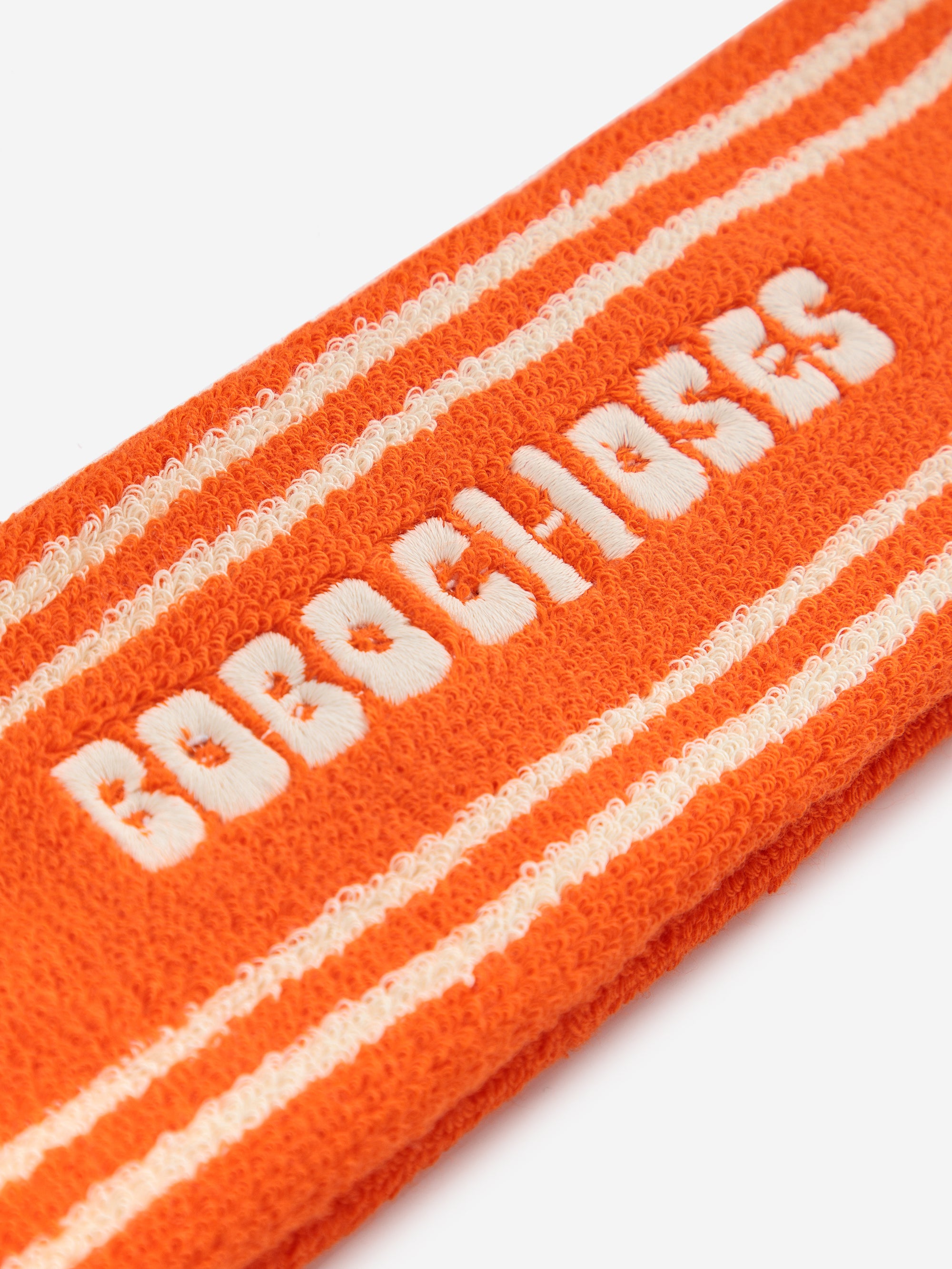 Close-up of Bobo Choses Orange Terry Towel Headband, showcasing its soft terry fabric, bold white embroidered logo, and sporty stripe accents.