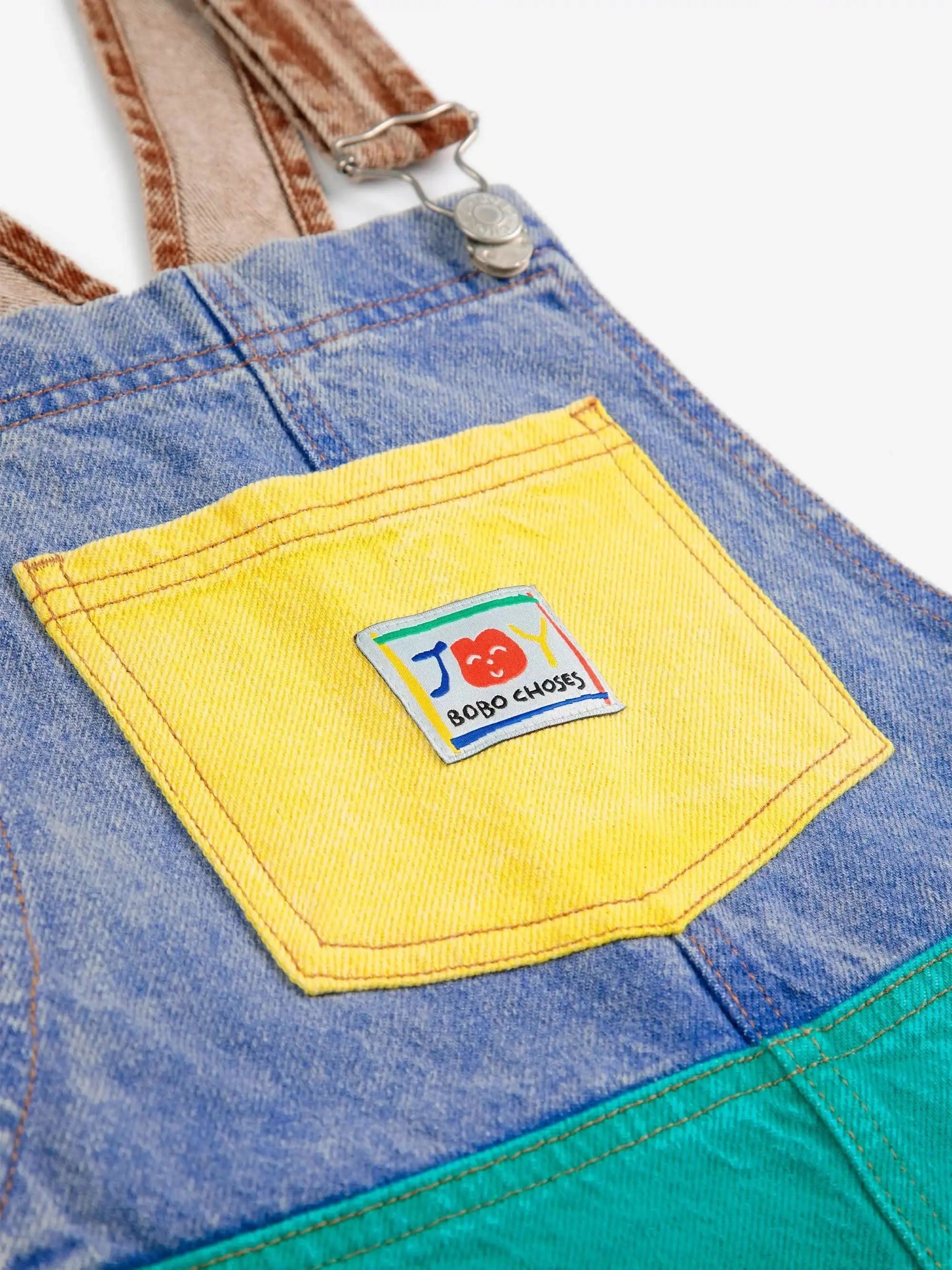 Detailansicht der gelben Brusttasche mit Bobo Choses Logo auf dem Smiling Color Block Denim Dungaree, trendiger Jumpsuit für Kinder.